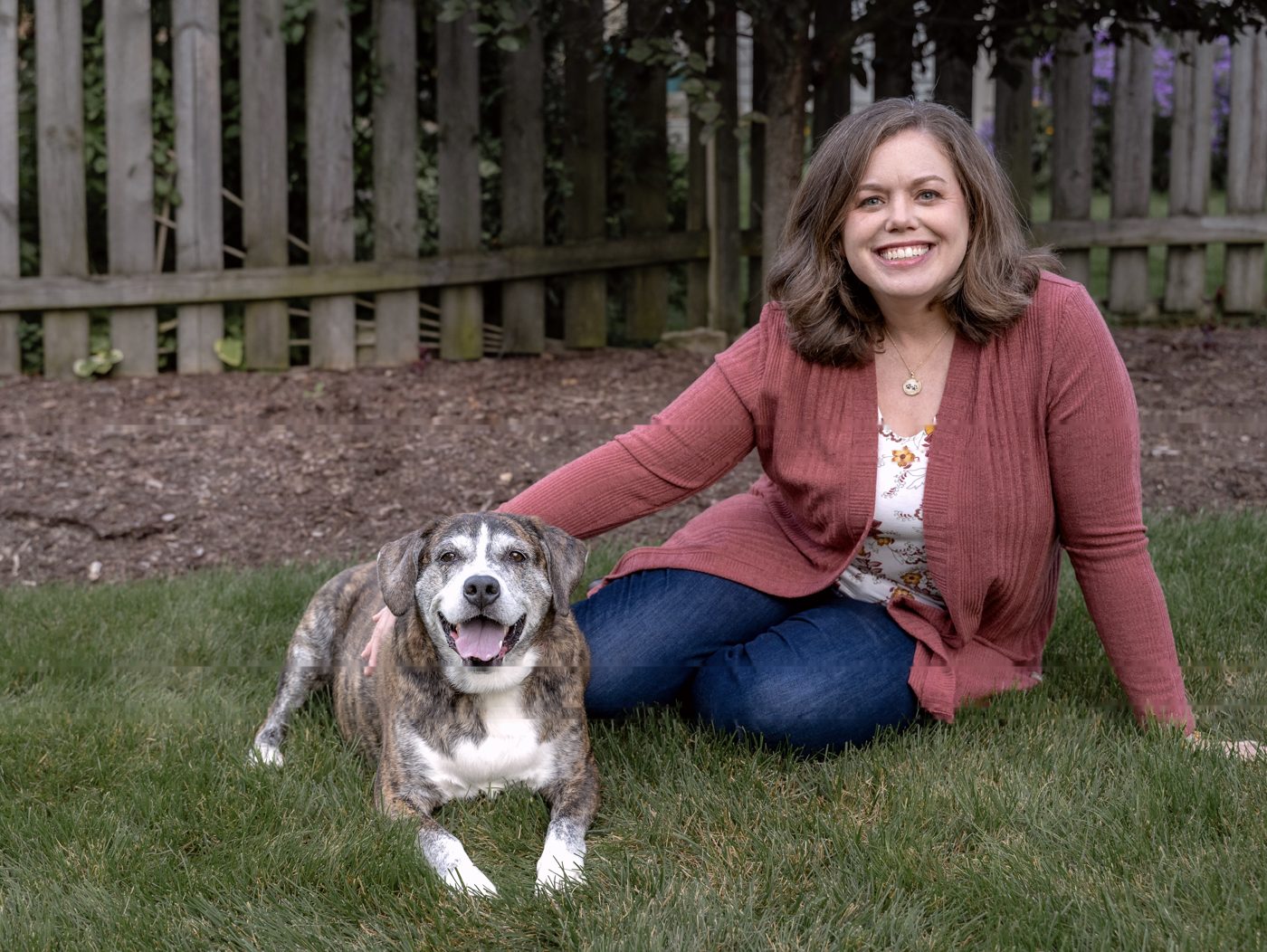 Dr. Christie sitting on the grass beside a smiling brindle dog